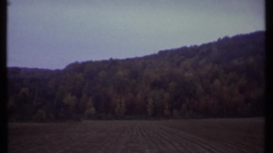 Rural Ithaca-area landscape from 1979 8mm footage, with a plowed field, wooded hillside, pale sky, and window edge in frame.