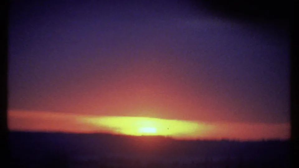 Soft 8mm frame of the Ithaca sunset, with an overexposed yellow sun band, dark hills, haze, and a small speck in the sky.