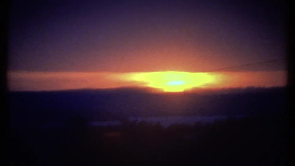 1979 Ithaca-area horizon at sunset, with a broad yellow-orange glow, silhouetted hills, dark foreground, and soft 8mm grain.