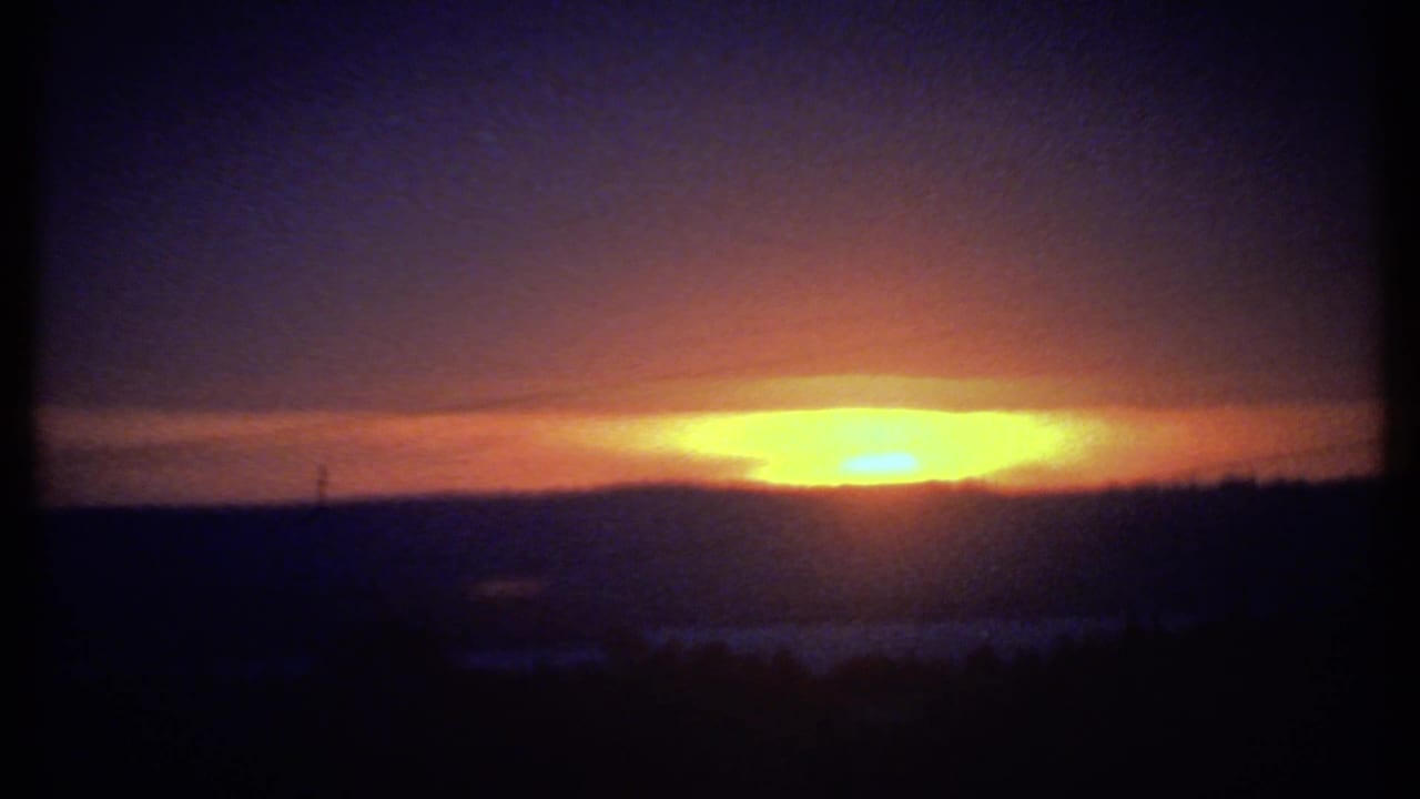 Likely Ithaca-area 1979 8mm frame of the sun setting behind hills, with dark foreground silhouettes and visible film grain.