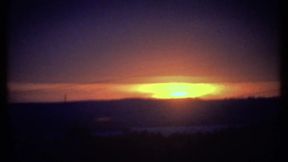 Likely Ithaca-area 1979 8mm frame of the sun setting behind hills, with dark foreground silhouettes and visible film grain.