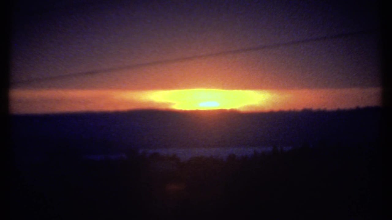 Bright sun sits near the horizon under a dark cloud layer with silhouetted landscape below.