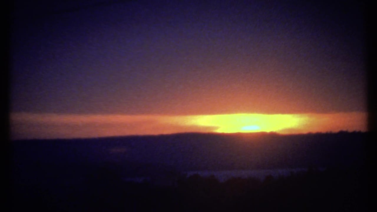 Orange sun near the horizon behind clouds above dark silhouetted hills.
