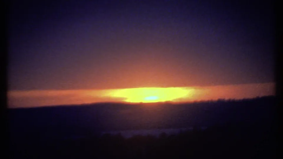 1979 8mm Ithaca frame showing the sun low behind a cloud bank, with saturated orange light fading into dark blue sky.