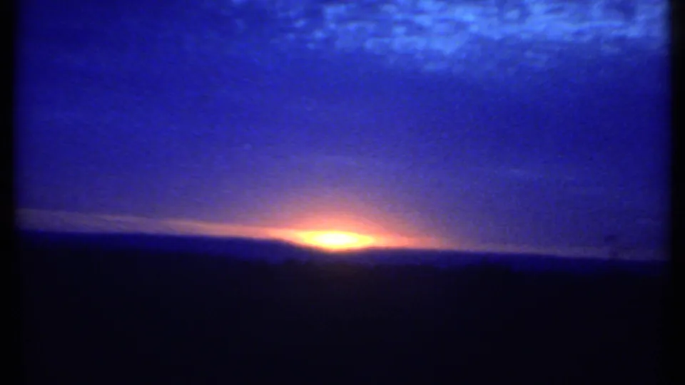 1979 8mm Ithaca sunset frame with a compact orange glow at the horizon under a dark blue sky and silhouetted land.