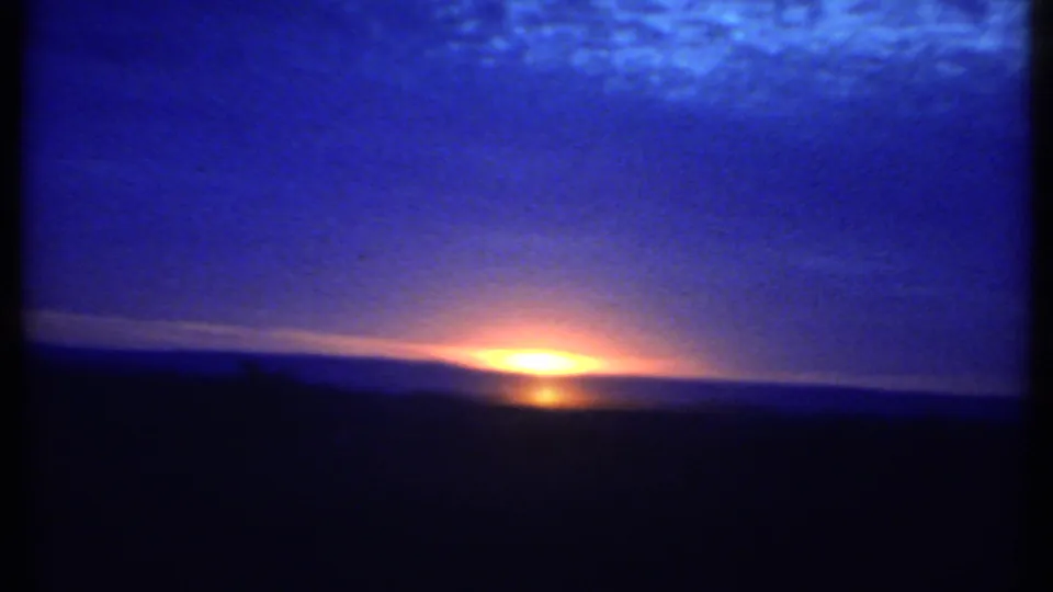 The low sun sits at the horizon in 1979 Ithaca 8mm footage, casting orange light beneath a grainy blue cloud bank.