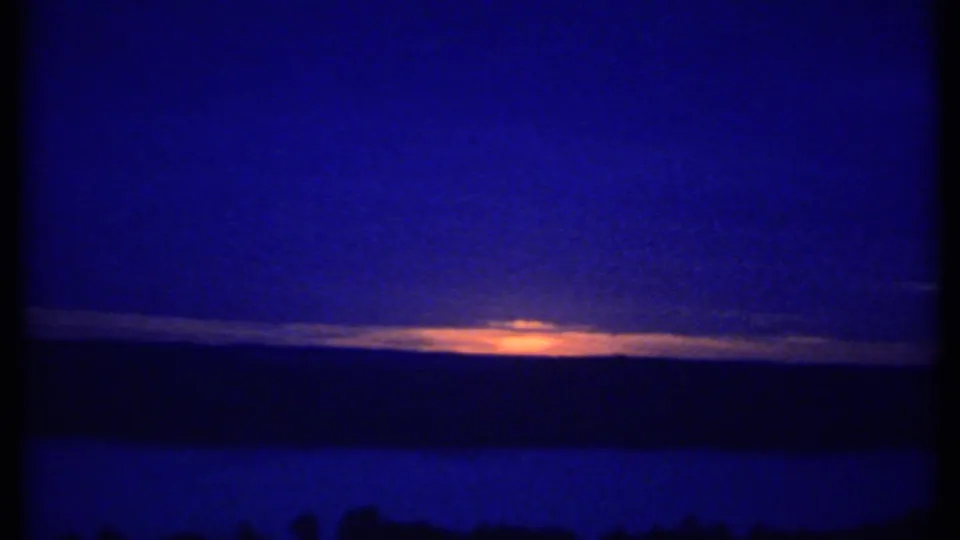 1979 8mm frame likely near Ithaca, New York, showing a warm sunset band behind clouds above a dark horizon and blue foreground.