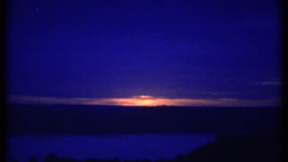 1979 8mm frame likely near Ithaca, New York, showing sunset light beneath clouds, a dark horizon, and a hillside silhouette at right.