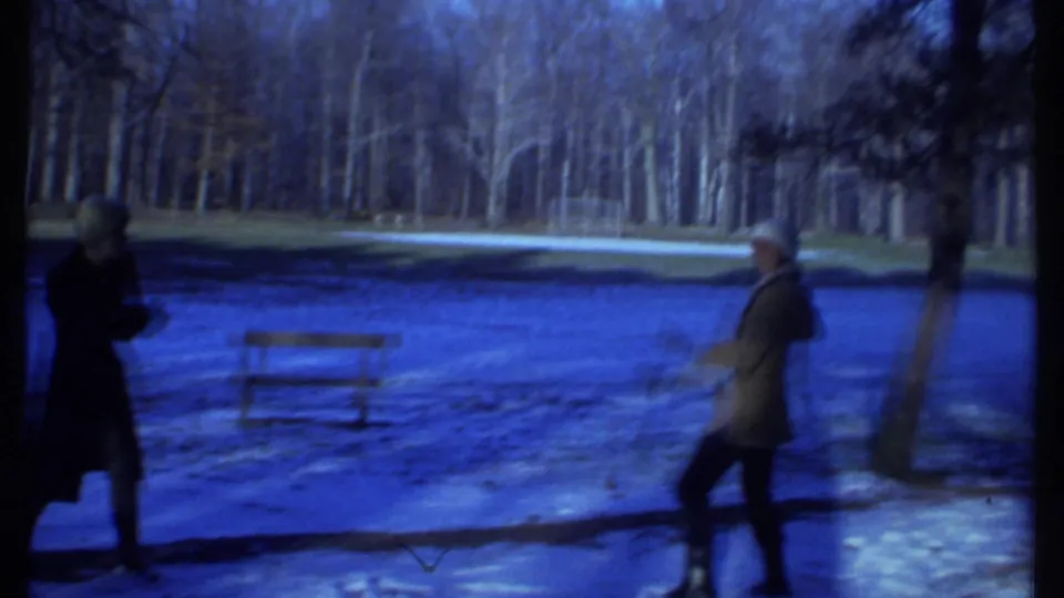 1979 Ithaca winter park scene with two distant bundled people, a bench, snow-covered ground, and bare trees.