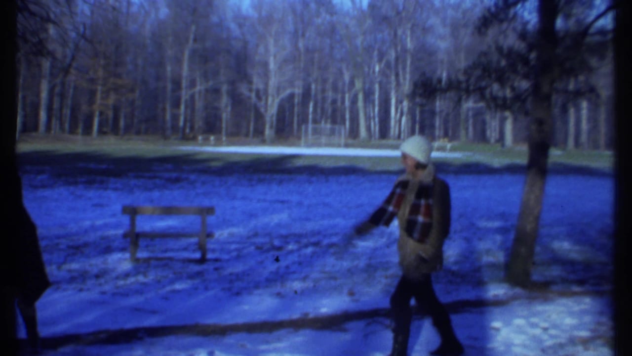 Bundled person in a white hat walks across snowy park ground near a wooden bench.