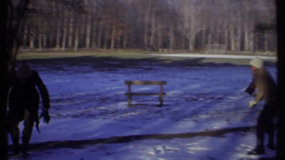 Two bundled people are visible in a snowy Ithaca park, one partly at the left edge and one moving near a bench.