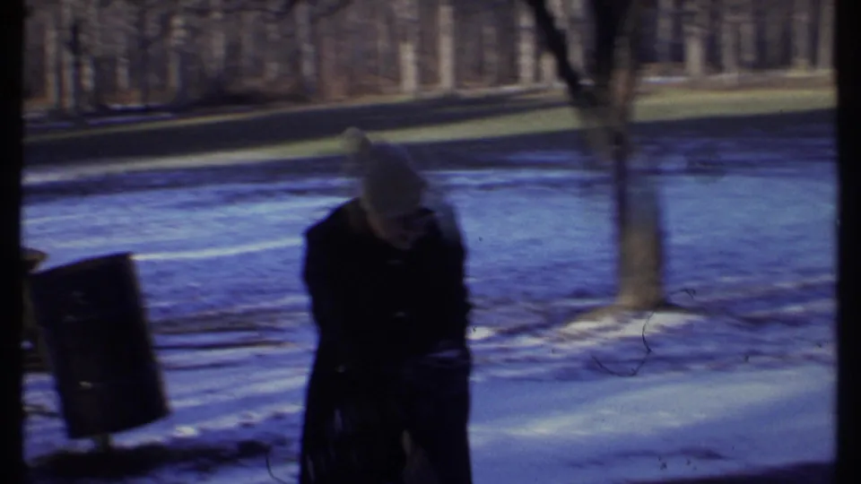 A bundled person in a knit hat stands in a snowy Ithaca park setting in 1979, with bare trees and a bench nearby.