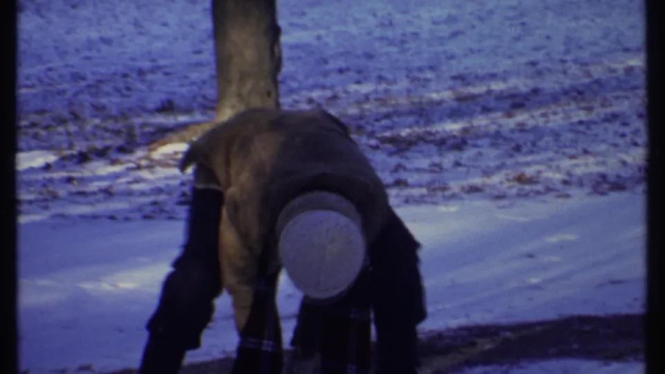 A bundled person bends down toward the snow near a bare tree in a winter park scene from Ithaca, New York, in 1979.
