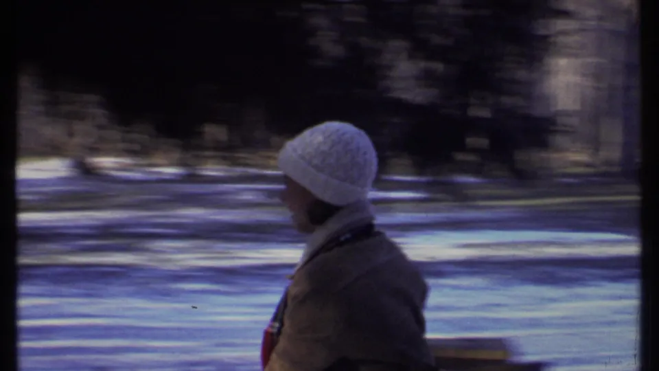 A person in a white knit hat moves through a snowy park area in Ithaca, with a bench and blurred trees visible behind them.