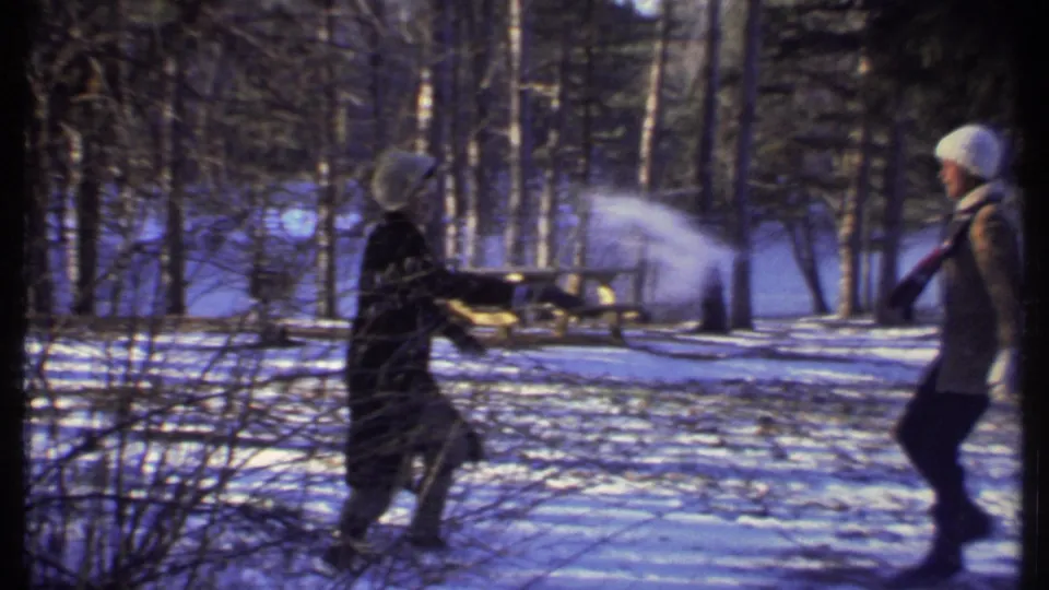 A person in dark winter clothing throws snow toward another bundled person in a snowy Ithaca park, with a picnic table behind them.
