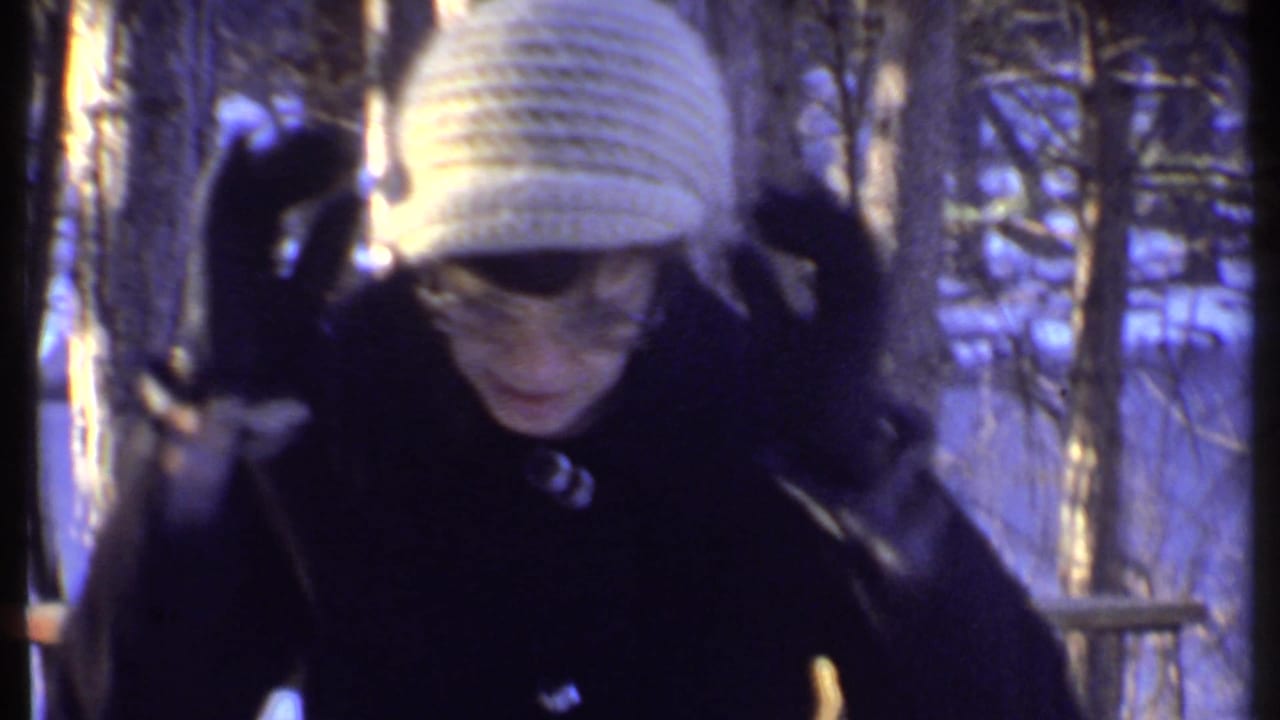 Woman in a white knit hat and dark coat adjusts her hat in a snowy Ithaca wooded setting.