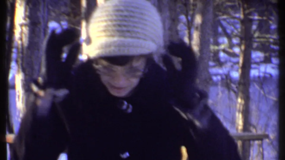 Woman in a white knit hat and dark coat adjusts her hat in a snowy Ithaca wooded setting.