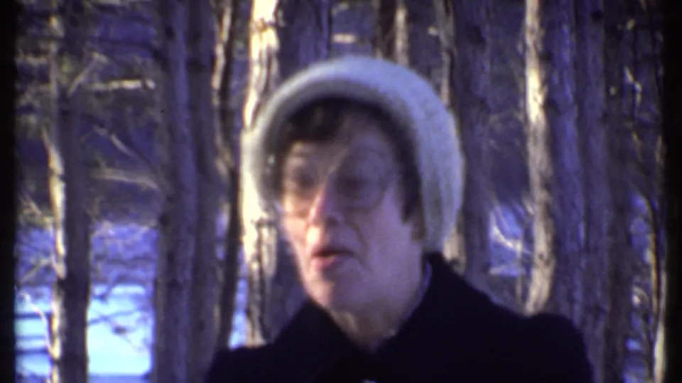 Woman in glasses, a white knit hat, and a dark coat faces the handheld 8mm camera near Ithaca in winter.