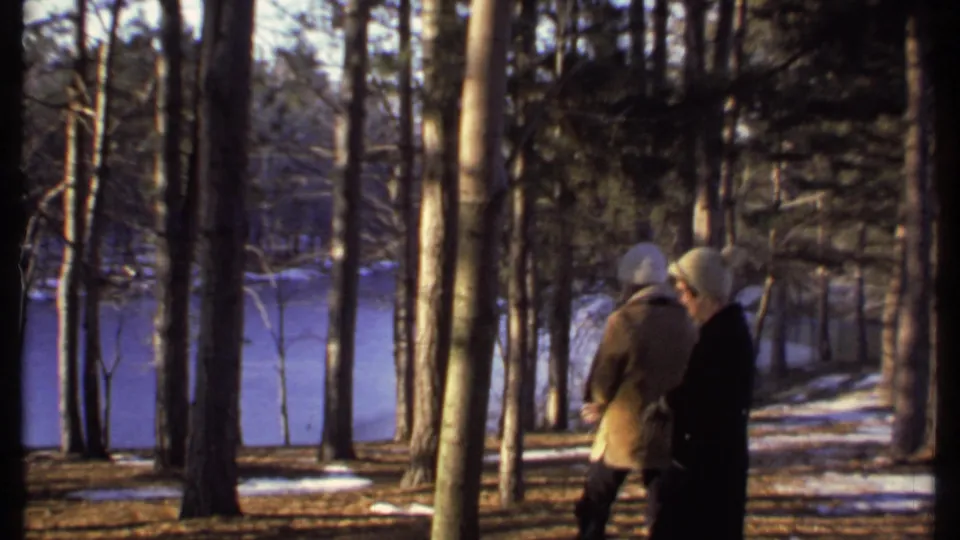 Two bundled people stand in a wooded area near a frozen lake in 1979 Ithaca, filmed in warm low winter light.