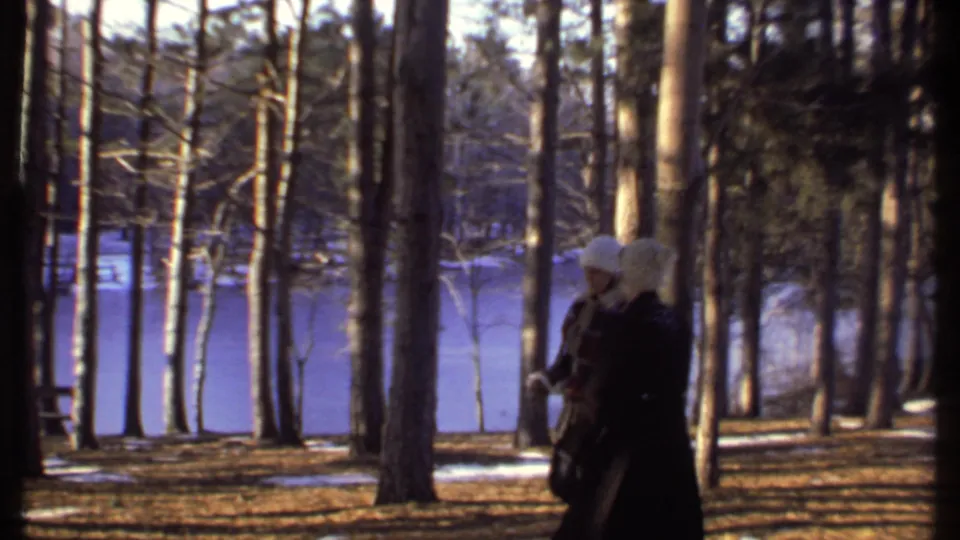 Two people in winter clothing walk together through trees near frozen water in the Ithaca home-movie footage.