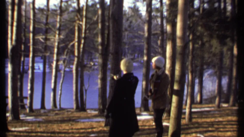 In 1979 Ithaca footage, one person in a dark winter coat gestures toward trees while another watches near the frozen lake.