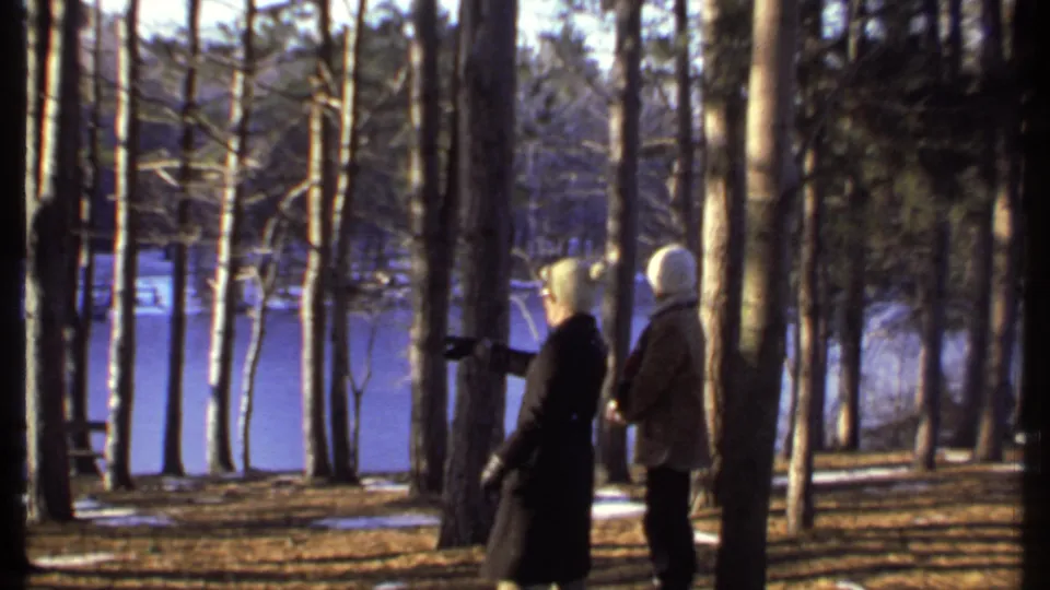 Two people in winter coats and white hats stand in profile among trees near a frozen lake in supported 1979 Ithaca footage.