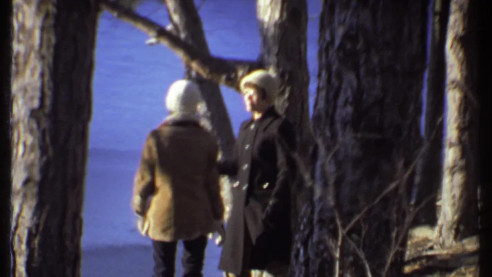 Two adults in winter coats and knit hats appear among trees during a 1979 Ithaca home-movie outing.