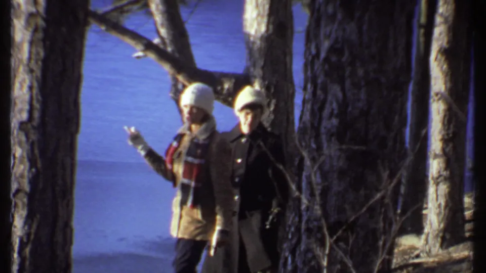 Two winter-dressed adults move through trees in Ithaca, New York, one raising a hand while walking.