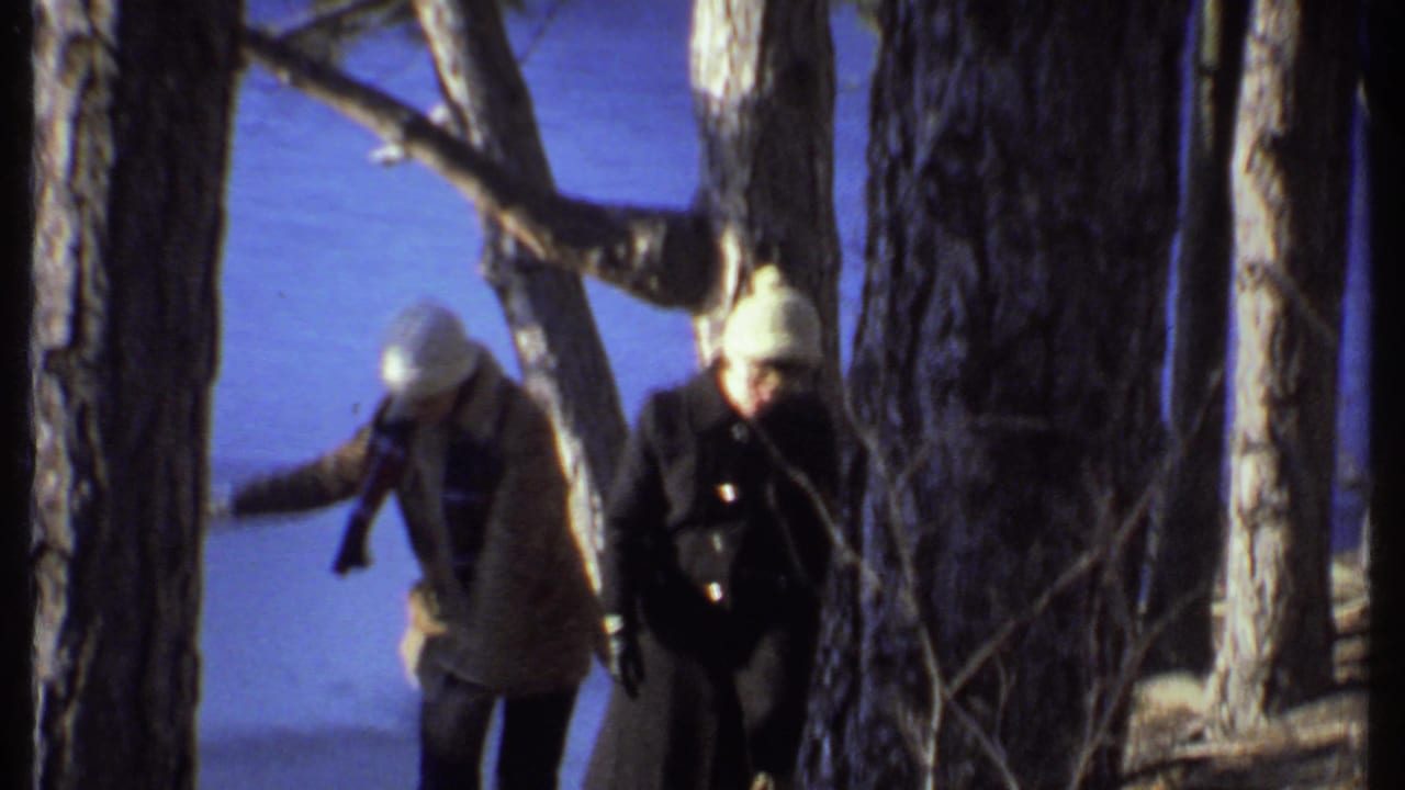 Two bundled adults walking among trees with blue lake water in the background.