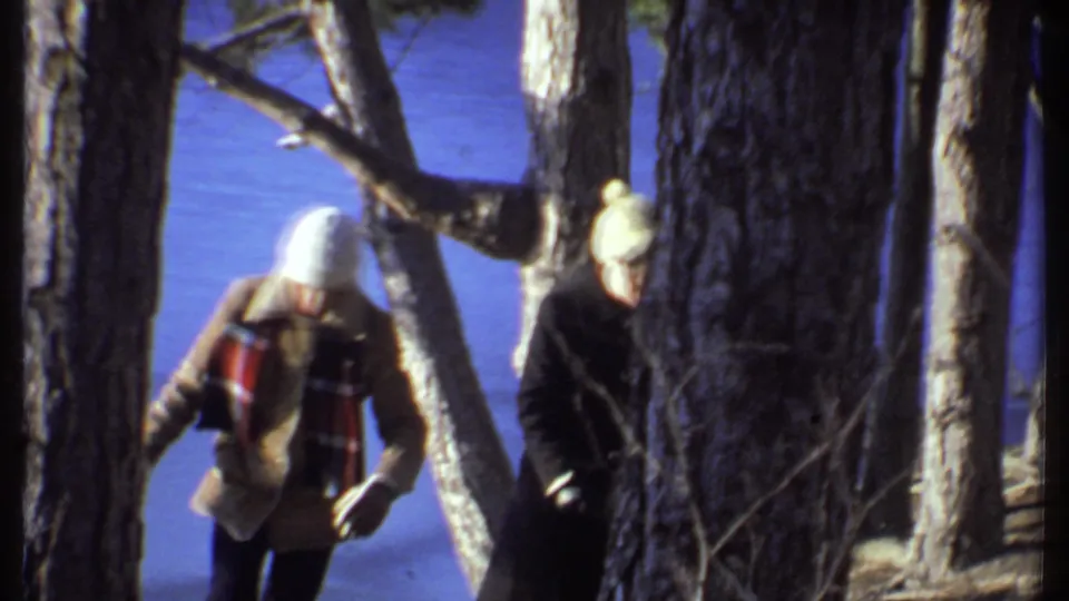 A bundled adult in a tan coat and plaid scarf walks past trees, with another adult in a dark coat nearby.