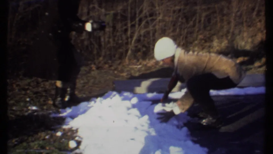 Two bundled people appear beside a snow pile in the 1979 Ithaca home movie, with one crouching to gather snow near a wooded pond edge.