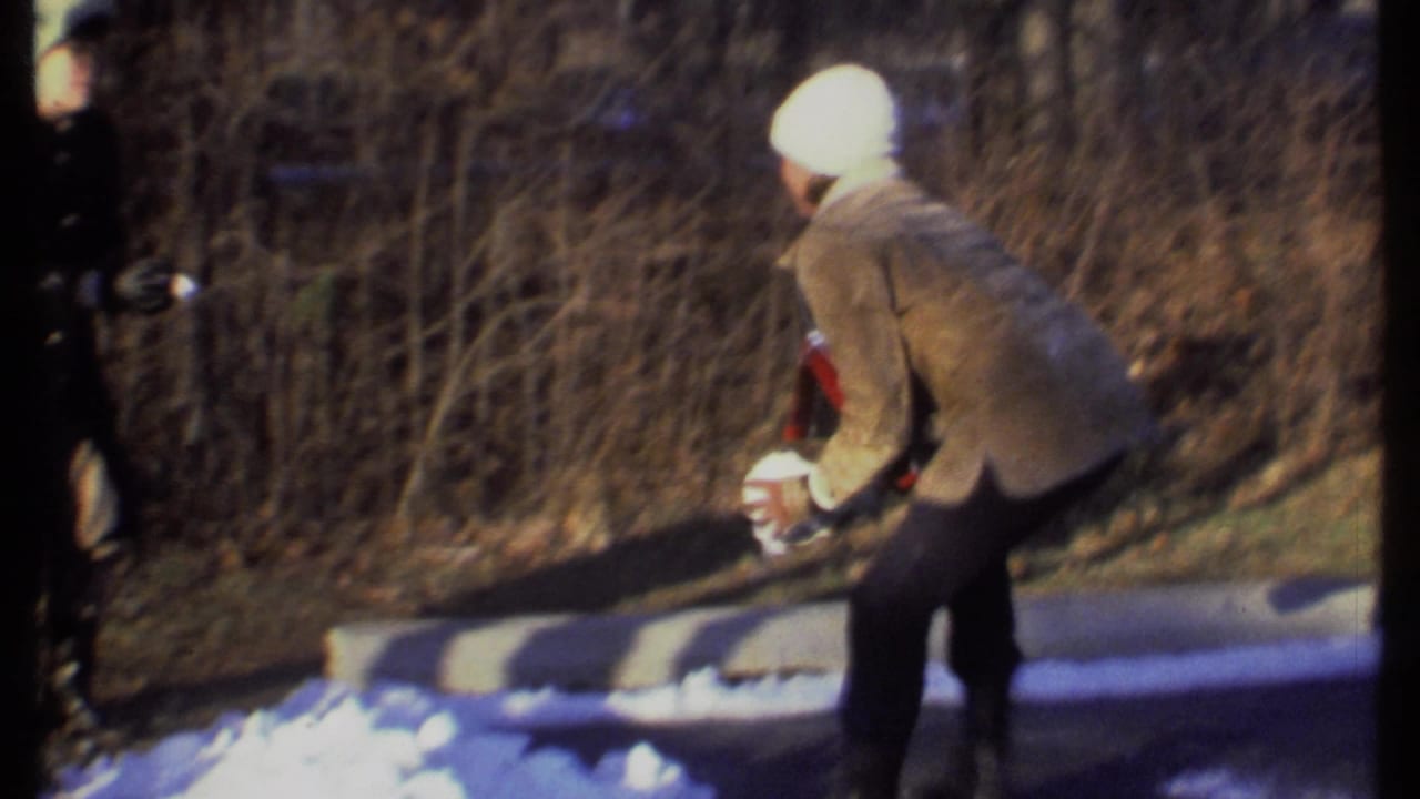 Person in a white knit hat bends near snow on a wooded path.