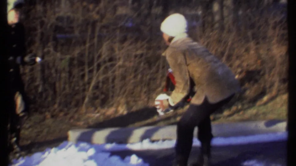 In Ithaca, New York, 1979 home-movie footage, a person in a white knit hat and tan coat bends to gather snow beside a wooded path.