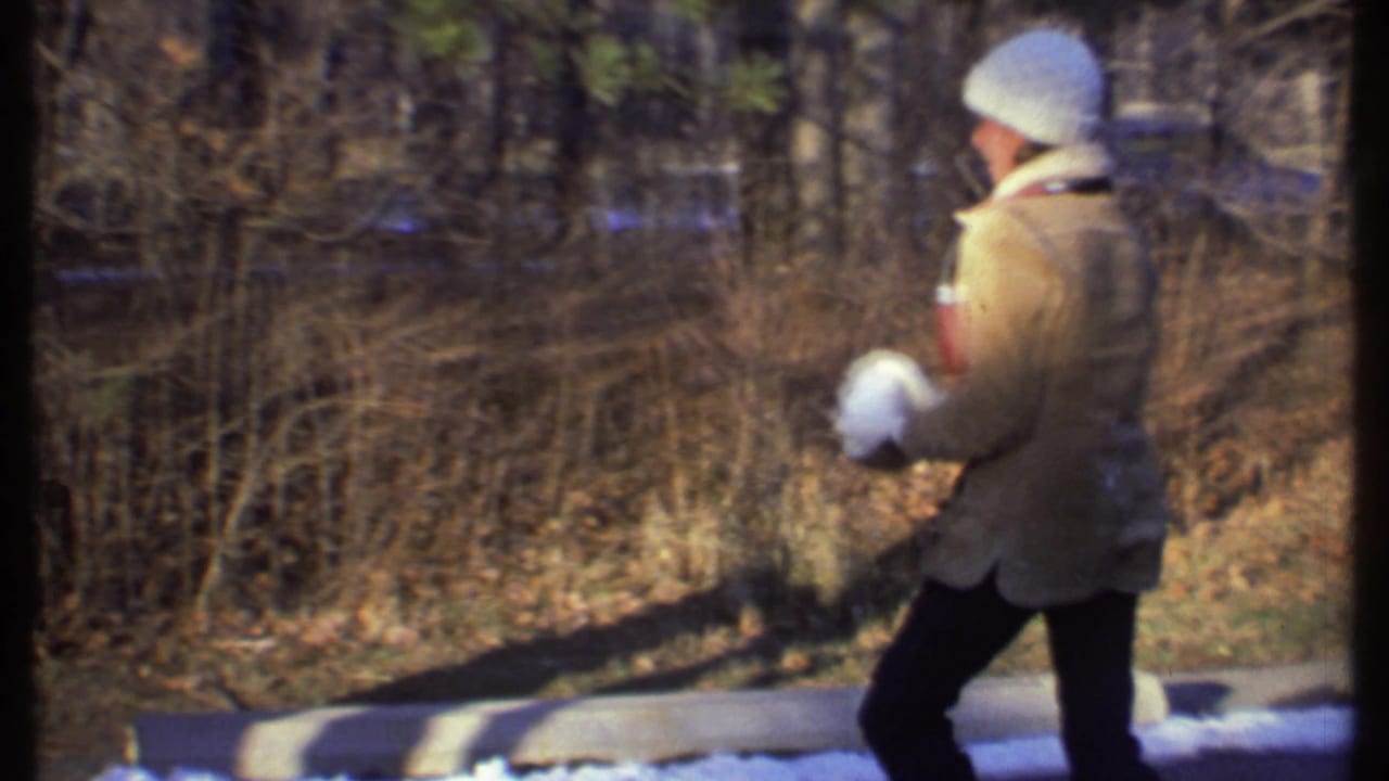 Person in a tan coat holds snowballs beside bare winter brush.