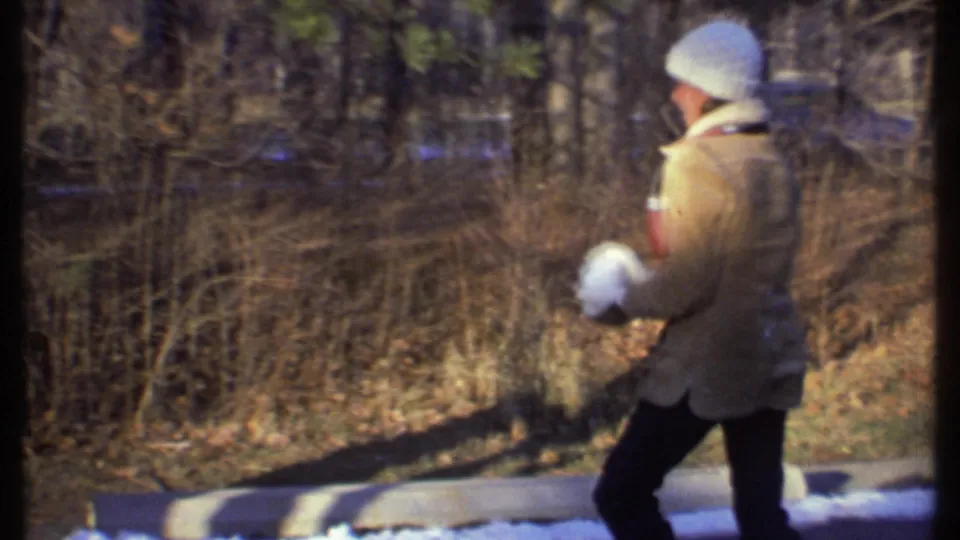A person in a tan coat and white knit hat holds snowballs near bare trees in the 1979 Ithaca winter scene.