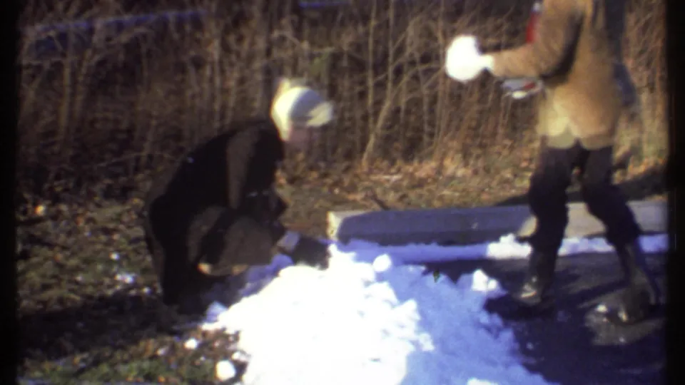 One person in dark clothing gathers snow while another figure in a tan coat stands at the edge of the snowy path in Ithaca, 1979.
