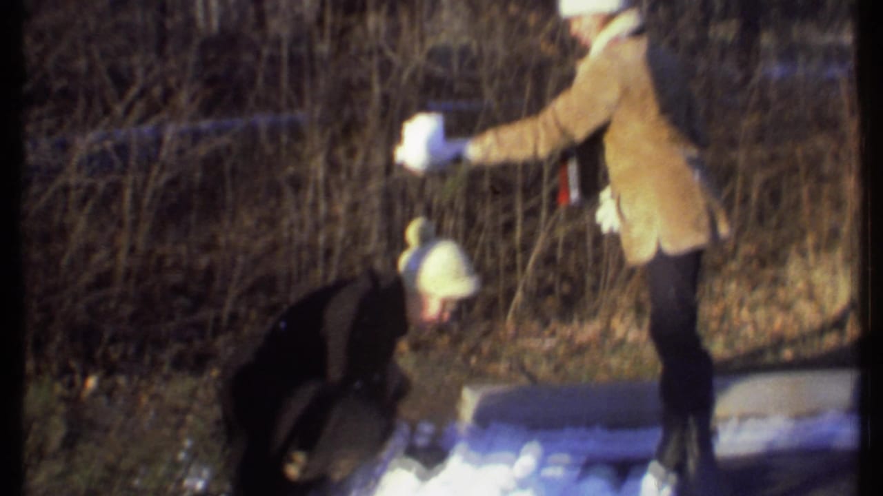 Two bundled people in snow near bare woods, one crouching while the other stands nearby.