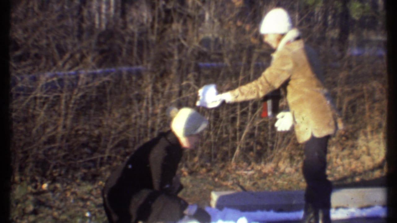 A crouching person gathers snow while another person in a tan coat holds a white object.