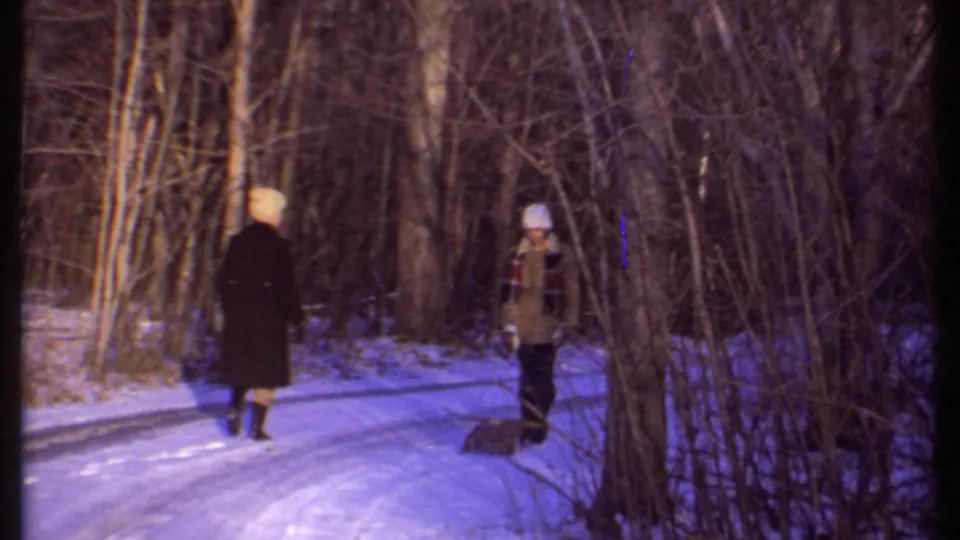 Two people in winter coats and hats stand on a snowy wooded path, with bare trees surrounding them in Ithaca.