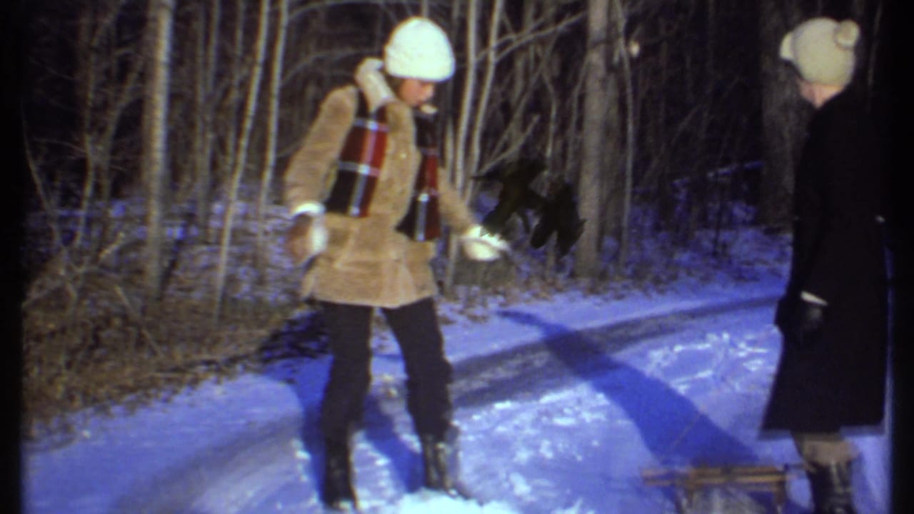 Two bundled adults stand on a snowy wooded path, one in a tan jacket and one in a black coat.