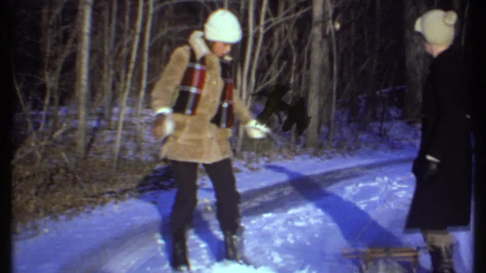 In 1979 Ithaca-area home-movie footage, two adults in winter coats and knit hats stand on a snowy path among bare trees.