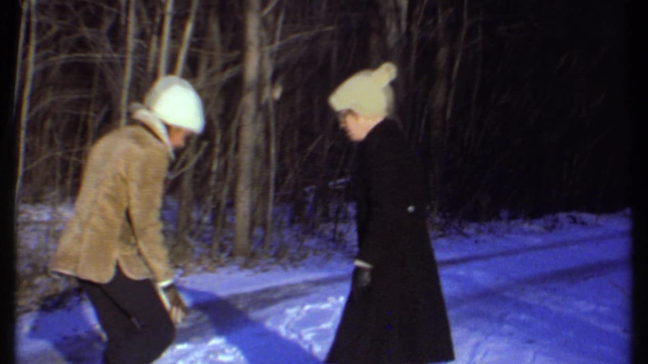 A bundled man bends toward the snow while a woman in a dark coat faces him on the path.