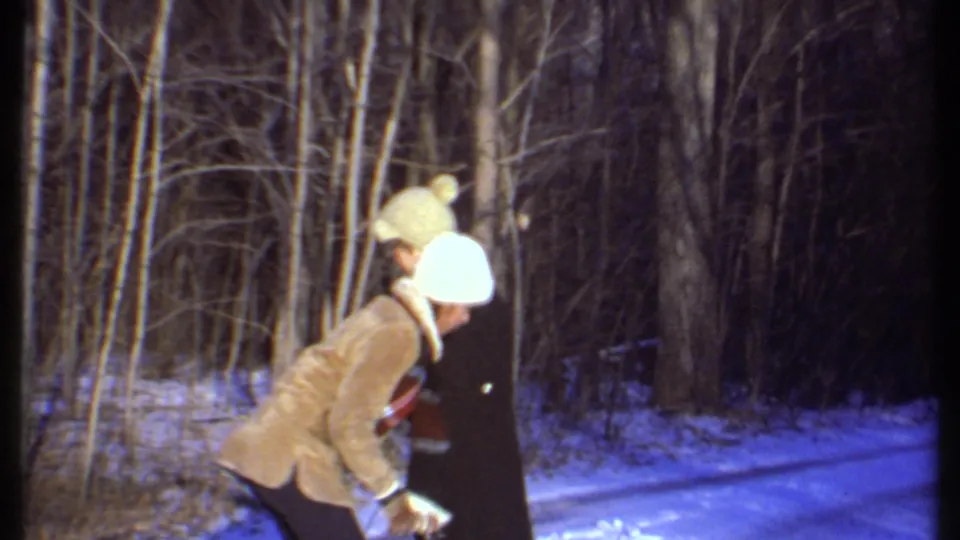 Two adults in winter clothing lean together on a snow-covered wooded path in 1979 home-movie footage from Ithaca.