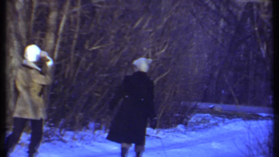 A woman in a tan coat appears to lift her arm during a playful snow moment as her companion walks ahead in the snowy woods.