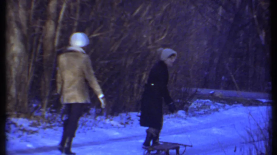 Two women in winter clothing continue through the snowy wooded setting, with a small sled or low object visible near the path.