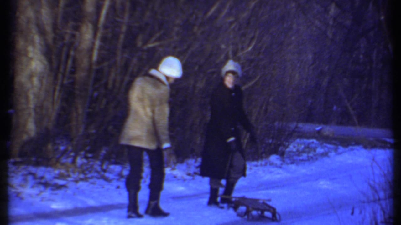 In Ithaca, New York, 1979, two people in winter coats and hats stand on a snow-covered path with a small sled nearby.
