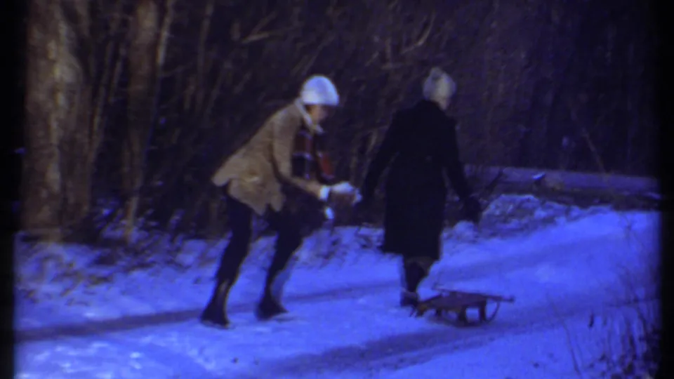A 1979 Ithaca winter home-movie frame shows one person reaching toward snow as another moves along the path with a small sled.