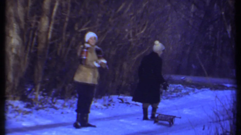 Two winter-clad people face each other on a snowy wooded path in Ithaca, with a small sled resting nearby.