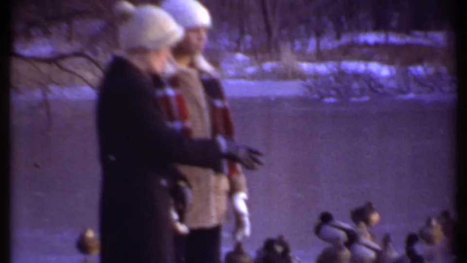 Two bundled figures stand by the frozen pond in the 1979 Ithaca home movie, facing ducks gathered on the icy surface.