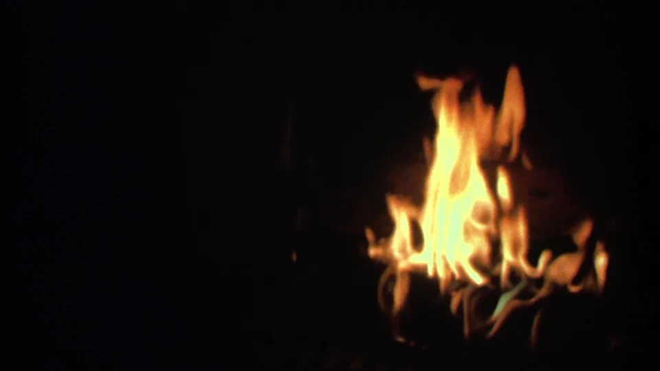 Tall orange-yellow flames rise in a dark night scene from 1979 Ithaca, New York 8mm footage, with grain and slight blur.